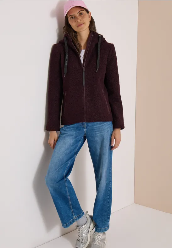 Frau in dunkler Kapuzenjacke, blauen Jeans und weißen Sneakers lehnt entspannt an heller Wand.