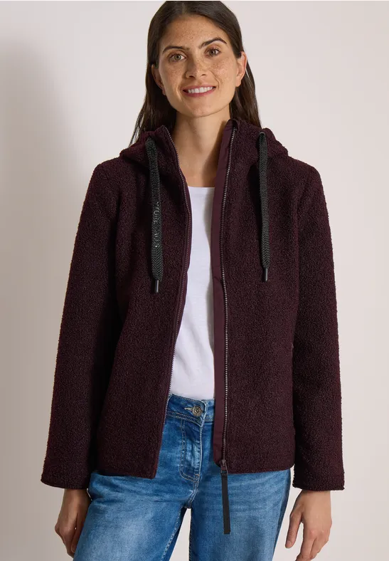 Junge Frau in jeans trägt eine flauschige, dunkelviolette Kapuzenjacke und lächelt.