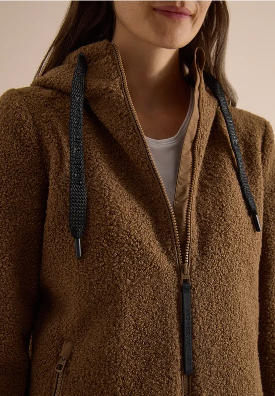 Braune Teddy-Fleece-Jacke mit Kapuze und Reißverschluss, darunter weißes Shirt.