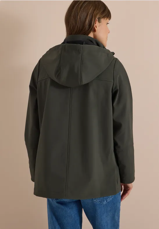 Une personne de dos portant une veste à capuche vert foncé et un jean devant un mur beige.