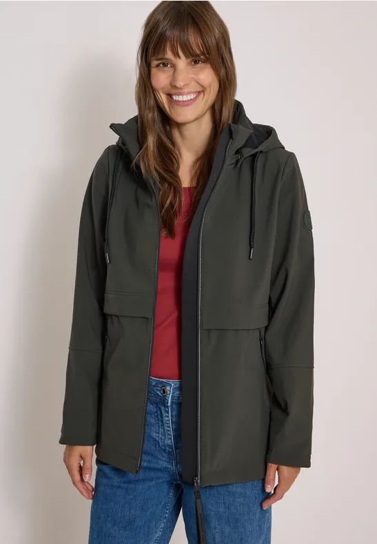 Une femme sourit, portant une veste verte sur un haut rouge et un jean devant un fond neutre