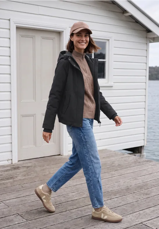 Frau mit schwarzer Jacke, beiger Mütze, braunem Pullover und Jeans steht lächelnd auf Holzsteg am Wasser.