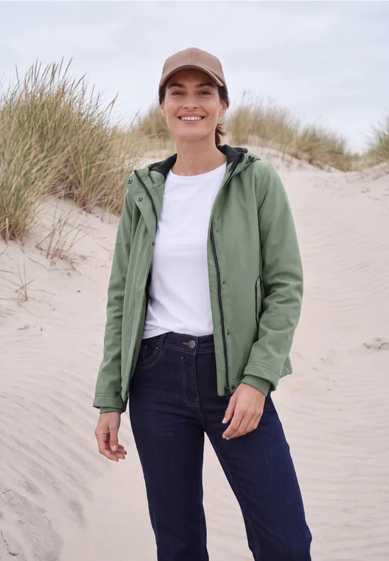 Eine lächelnde Frau in weißem Shirt, grüner Jacke, Jeans und Cap steht am Dünenstrand.