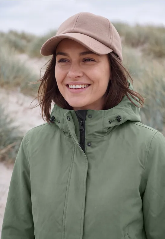 Lächelnde Frau mit grüner Jacke und beiger Baseballkappe vor Dünenkulisse.