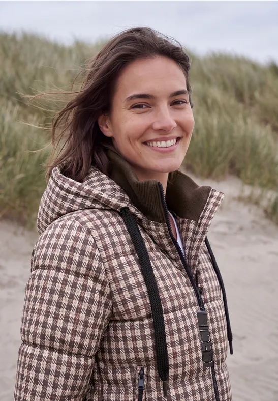 Eine lächelnde junge Frau in kariertem Mantel steht vor Dünen am Strand.