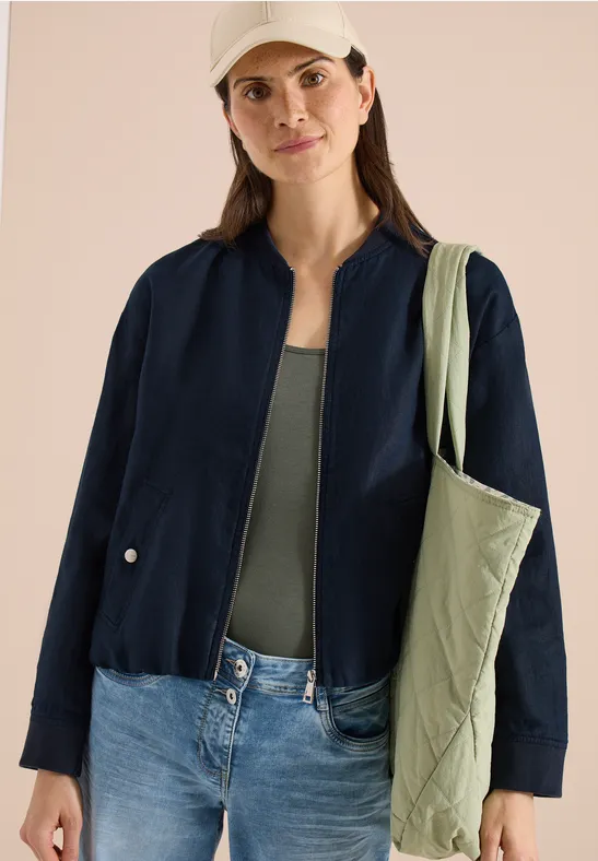 Frau in dunkler Jacke, grünem Top, Jeans und hellgrüner Stepp-Tasche über der Schulter.