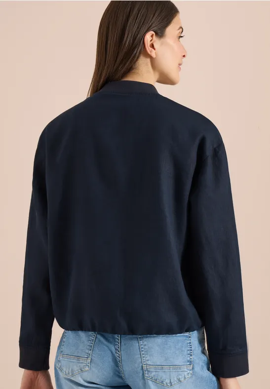 Frau von hinten in dunkelblauem Cardigan und Jeans vor hellem Hintergrund.