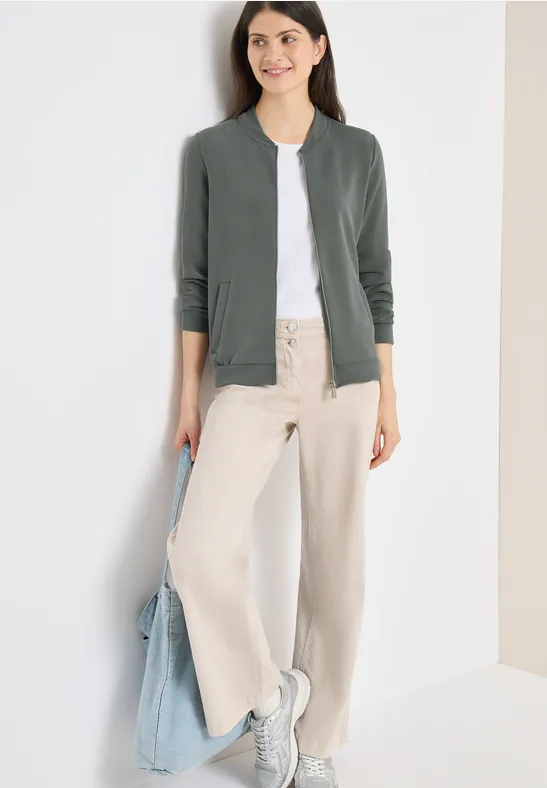 Frau trägt graugrünen Cardigan, weißes T-Shirt, weite beiger Hose, weiße Sneaker und hält eine hellblaue Tasche.