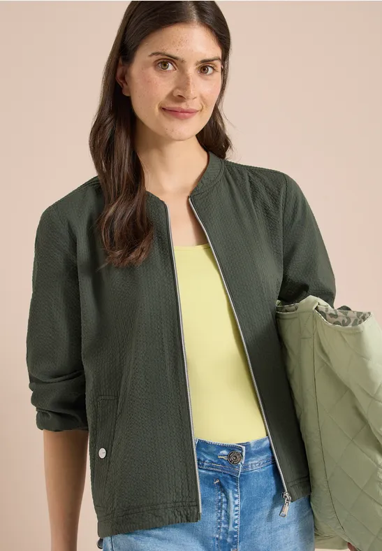 Vrouw in donkergroene jas, geel topje en spijkerbroek, met een groene tas in de hand.