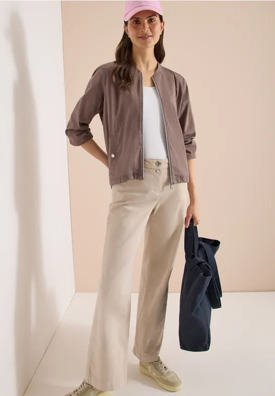 Persoon draagt witte top, lila blouse, lichte wijde broek, groene sneakers en houdt een blauwe jas.