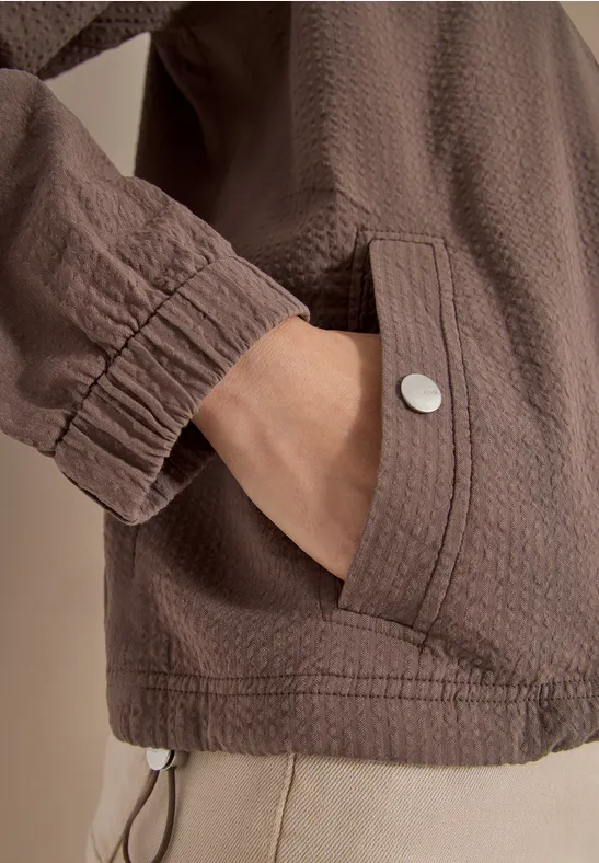 Bruin gestructureerd jasje met opgestikte zak en metalen knoop, hand in de zak.