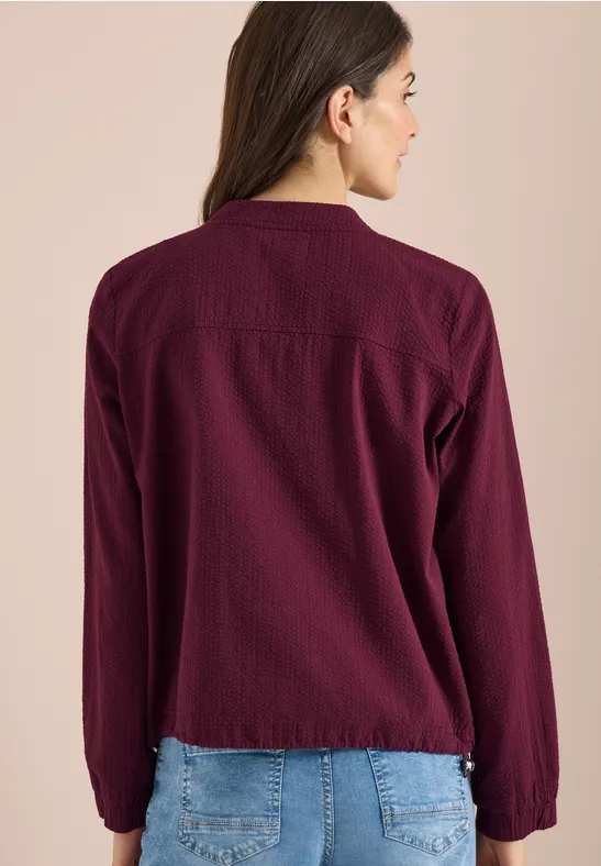 Frau von hinten in bordeauxroter Bluse und blauer Jeans vor beigem Hintergrund.