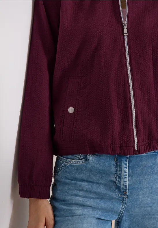 Bordeauxroter Reißverschluss-Blouson mit Strukturstoff und aufgesetzten Taschen, kombiniert mit blauen Jeans.