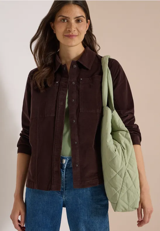 Femme en veste en velours côtelé brune, chemisier vert, jean bleu et grand sac matelassé vert.