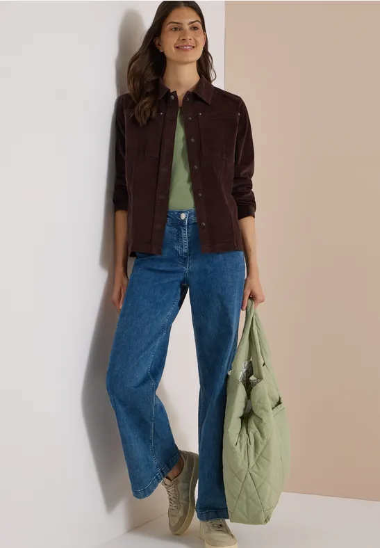 Femme en jean ample, T-shirt vert, veste en velours marron, tenant un grand sac vert matelassé.