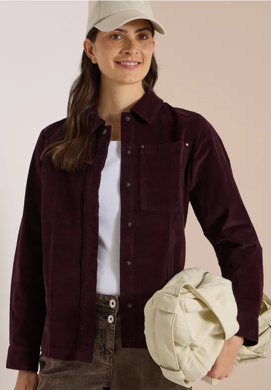 Femme souriante en veste en velours côtelé bordeaux, t-shirt blanc et pantalon vert, tenant un sac crème.
