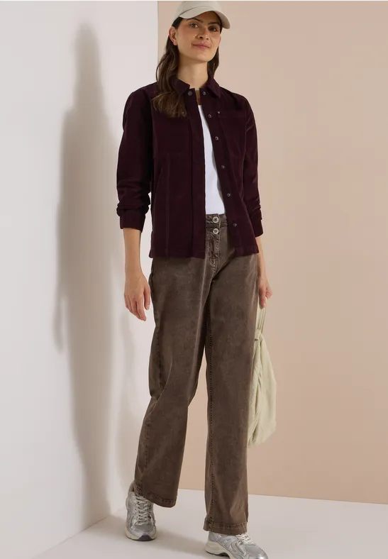 Femme en look décontracté : top blanc, veste bordeaux, pantalon velours marron, baskets argentées, casquette.