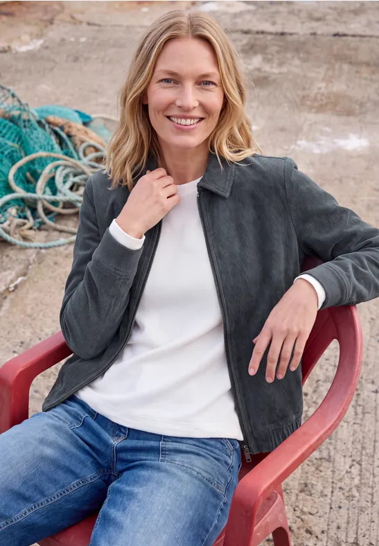 Eine Frau mit blonden Haaren sitzt draußen auf einem roten Stuhl und lächelt, trägt Jeans und Jacke.