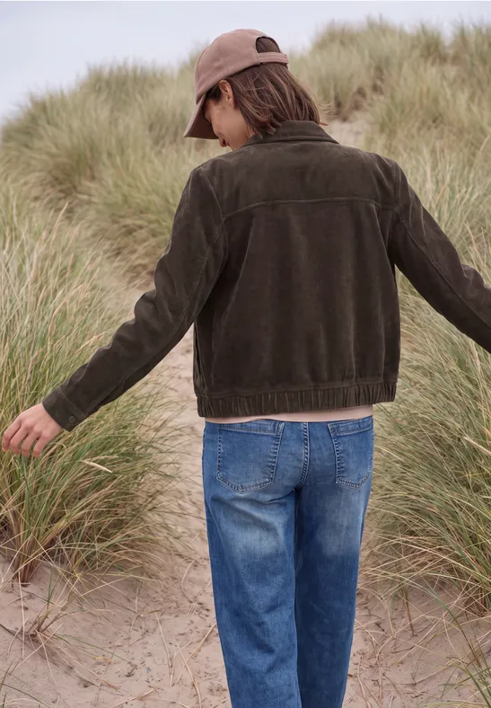 Rückansicht einer Person in Jeans und dunkler Jacke, die durch Gras auf sandigen Dünen geht.
