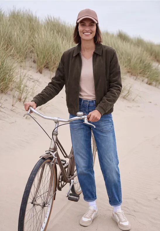 Eine Frau mit Fahrrad steht lächelnd auf einem Sanddünenstrand, trägt Jeans und Jacke.
