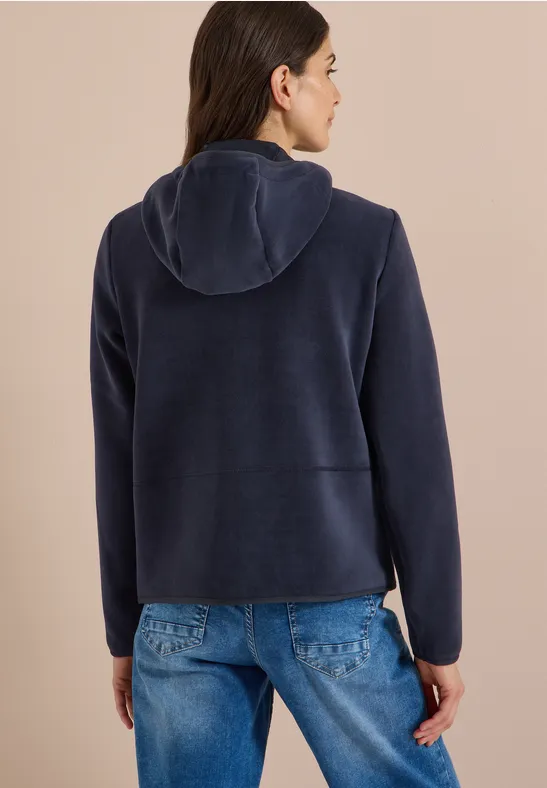 Frau von hinten trägt dunkelblauen Kapuzen-Fleece und Jeans vor hellem Hintergrund.