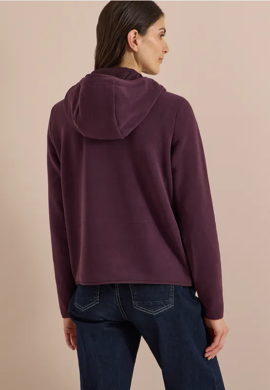 Rückansicht einer Person in dunkelviolettem Kapuzenpullover und blauer Jeans vor beigem Hintergrund.