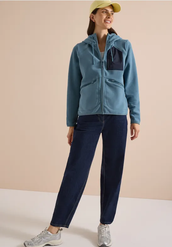 Vrouw glimlacht in blauwe fleecejas, donkerblauwe broek, lichte pet en sneakers tegen een neutrale achtergrond.