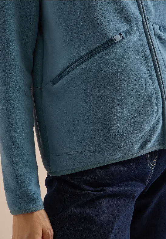 Persoon draagt turquoise fleecevest met ritssluiting en blauwe spijkerbroek.