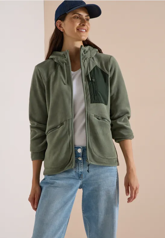 Vrouw met groene fleecevest met capuchon, witte top, spijkerbroek en pet.