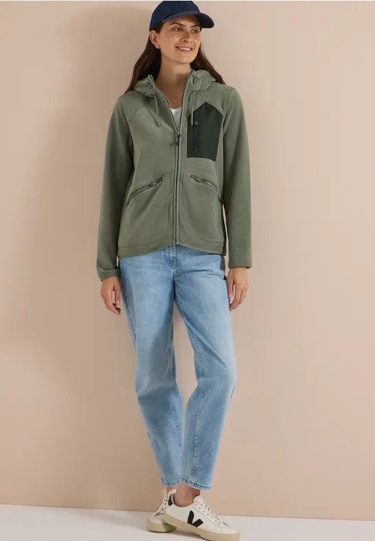 Vrouw in groene jas, spijkerbroek, sneakers en pet voor een beige achtergrond.