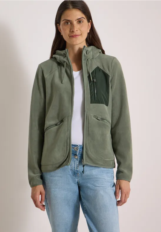 Een vrouw staat frontaal, draagt een groene fleecevest met kap, wit shirt en lichtblauwe spijkerbroek.