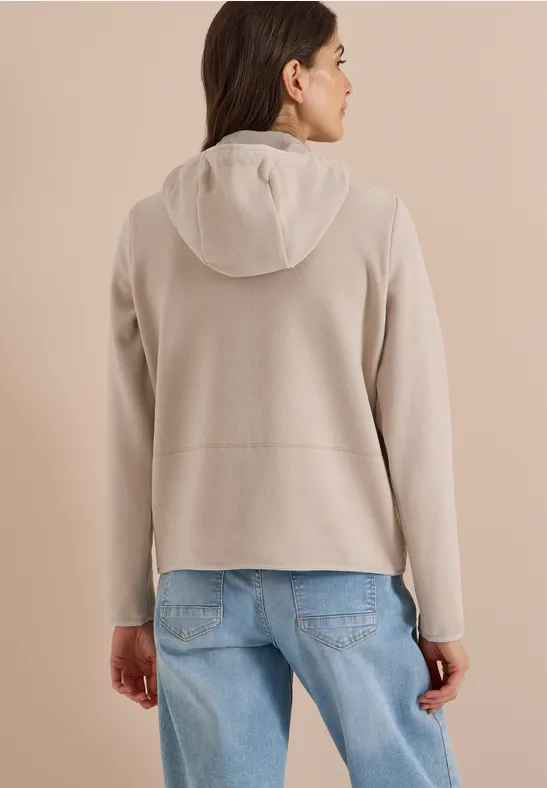 Frau von hinten in hellgrauer Fleece-Jacke mit Kapuze und blauen Jeans vor neutralem Hintergrund.