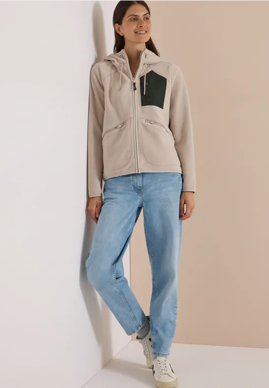 Frau lehnt an Wand in Freizeit-Outfit mit beiger Fleecejacke, Jeans und weißen Sneakern.