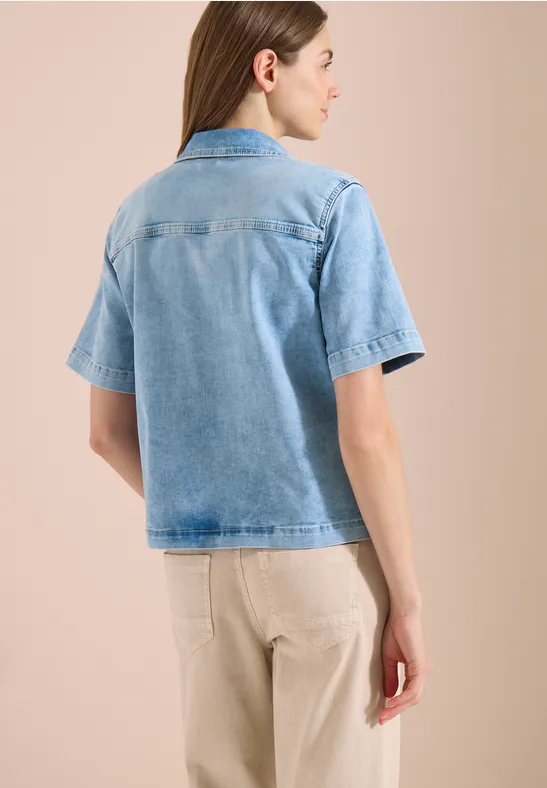 Frau in blauem Jeans-Hemd mit kurzen Ärmeln und beiger Hose, Rückenansicht vor neutralem Hintergrund.