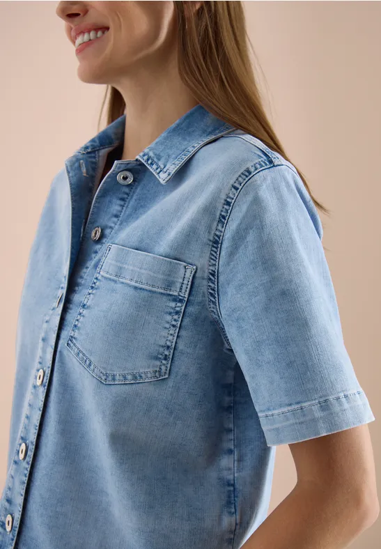 Frau trägt hellblaue Jeansbluse mit kurzen Ärmeln und Brusttasche vor neutralem Hintergrund.