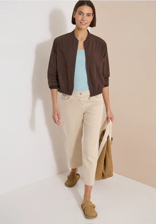 Femme en bomber marron, top bleu clair, pantalon beige, mocassins verts et sac en osier sur fond clair.