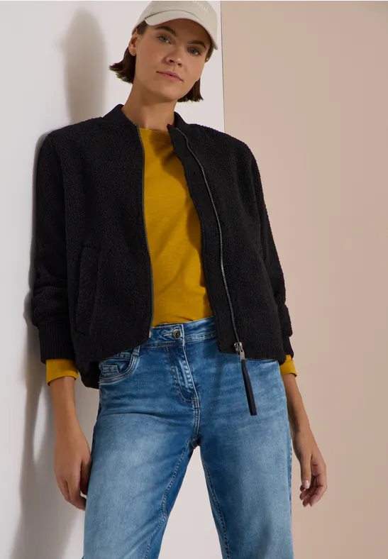 Vrouw tegen muur, zwarte jas, gele trui, blauwe jeans en witte pet.