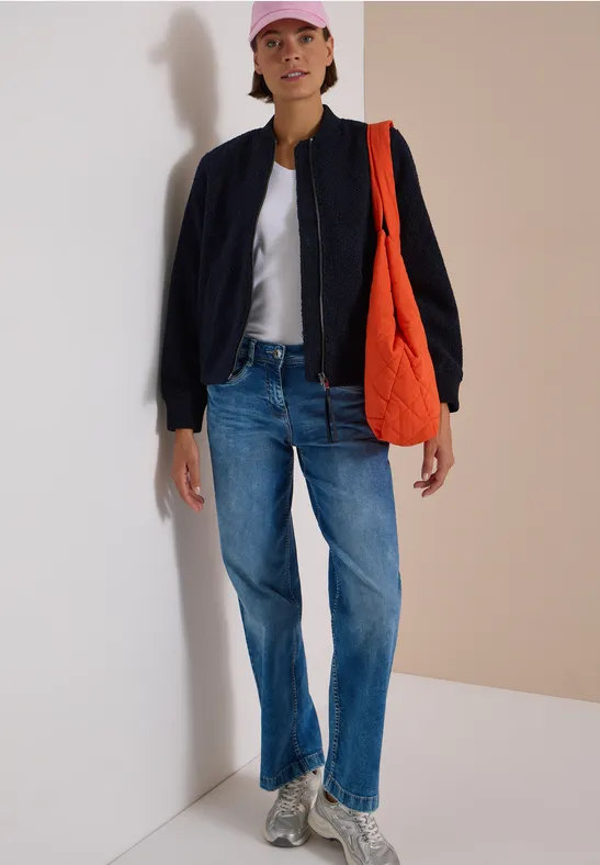 Iemand in een casual look: jeans, wit shirt, zwarte jas, roze pet, grijze sneakers en een oranje tas voor een lichte muur.