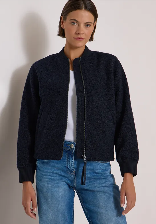 Vrouw met donkerblauw fleece bomberjack, wit T-shirt en blauwe spijkerbroek voor een neutrale achtergrond.