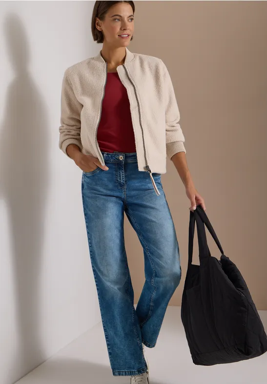 Frau in Bomberjacke, rotem Shirt, Jeans und großer Tasche lehnt lässig an weißer Wand.