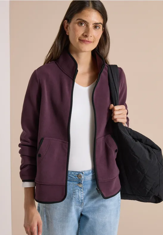 Vrouw draagt paarse jas, wit topje, spijkerbroek en houdt een zwarte tas.