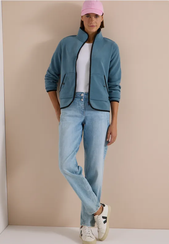 Eine Frau steht in Jeans, weißem Shirt, blauem Fleece, rosa Cap und Sneakern vor beige.