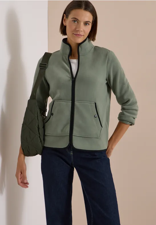 Frau trägt grüne Fleecejacke, weißes Shirt, blaue Jeans und grüne Umhängetasche vor hellem Hintergrund.