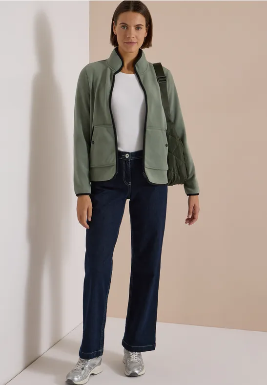 Frau steht in Raumecke, trägt dunkle Jeans, weiße Bluse, grüne Jacke und Sneaker.