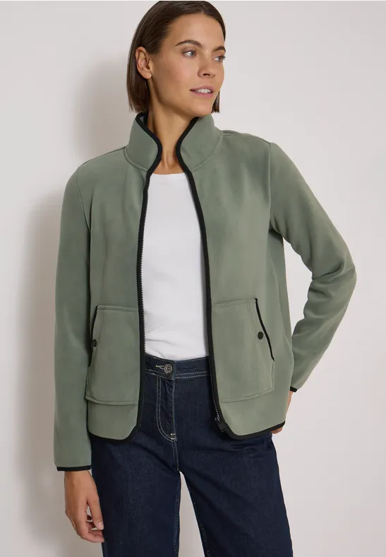 Frau trägt olivgrüne Fleecejacke mit Reißverschluss und Taschen, kombiniert mit weißem Shirt und Jeans.