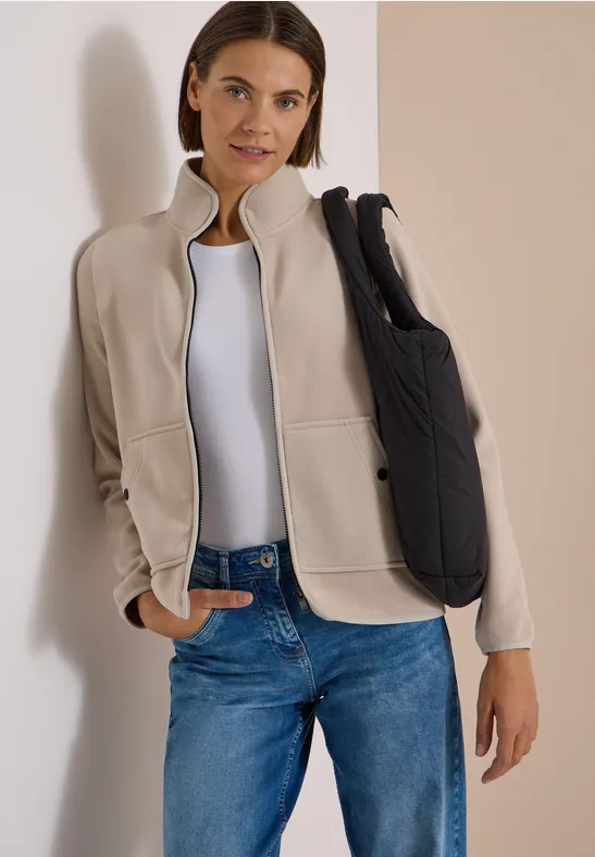 Frau in beiger Jacke, weißem T-Shirt und blauen Jeans mit schwarzer Umhängetasche.