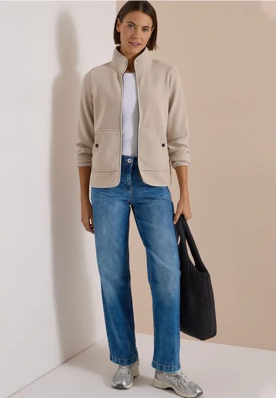Frau in heller Jacke, weißem Shirt, blauen Jeans und silbernen Sneakern mit schwarzer Tasche vor neutraler Wand.