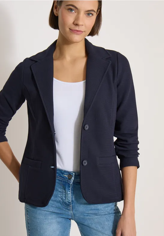Une femme porte un blazer bleu marine, un haut blanc et un jean sur fond clair.