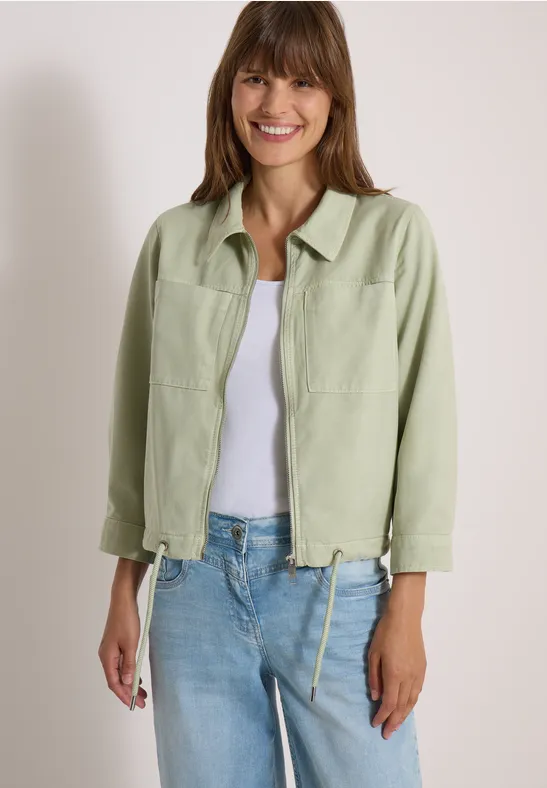 Eine lächelnde Frau in hellgrüner Jacke, weißem Top und Jeans vor neutralem Hintergrund.