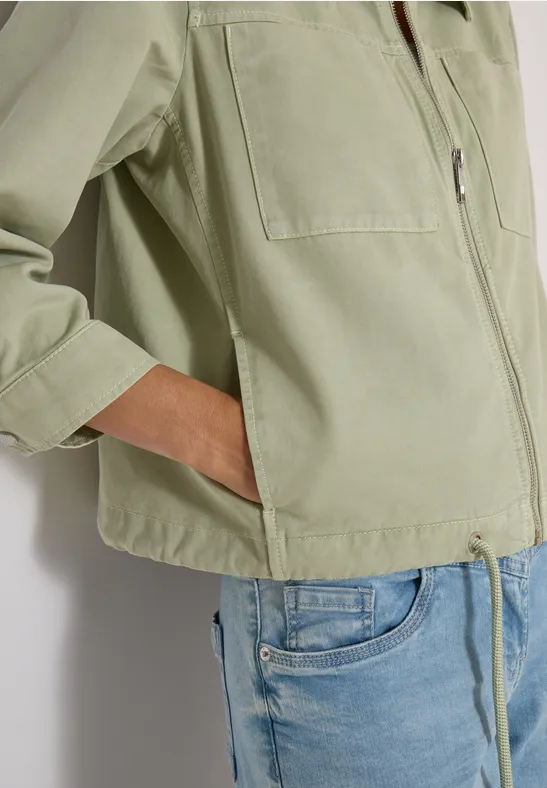 Person trägt eine helle, olivgrüne Jacke mit eingesteckter Hand in der Tasche und hellblaue Jeans.